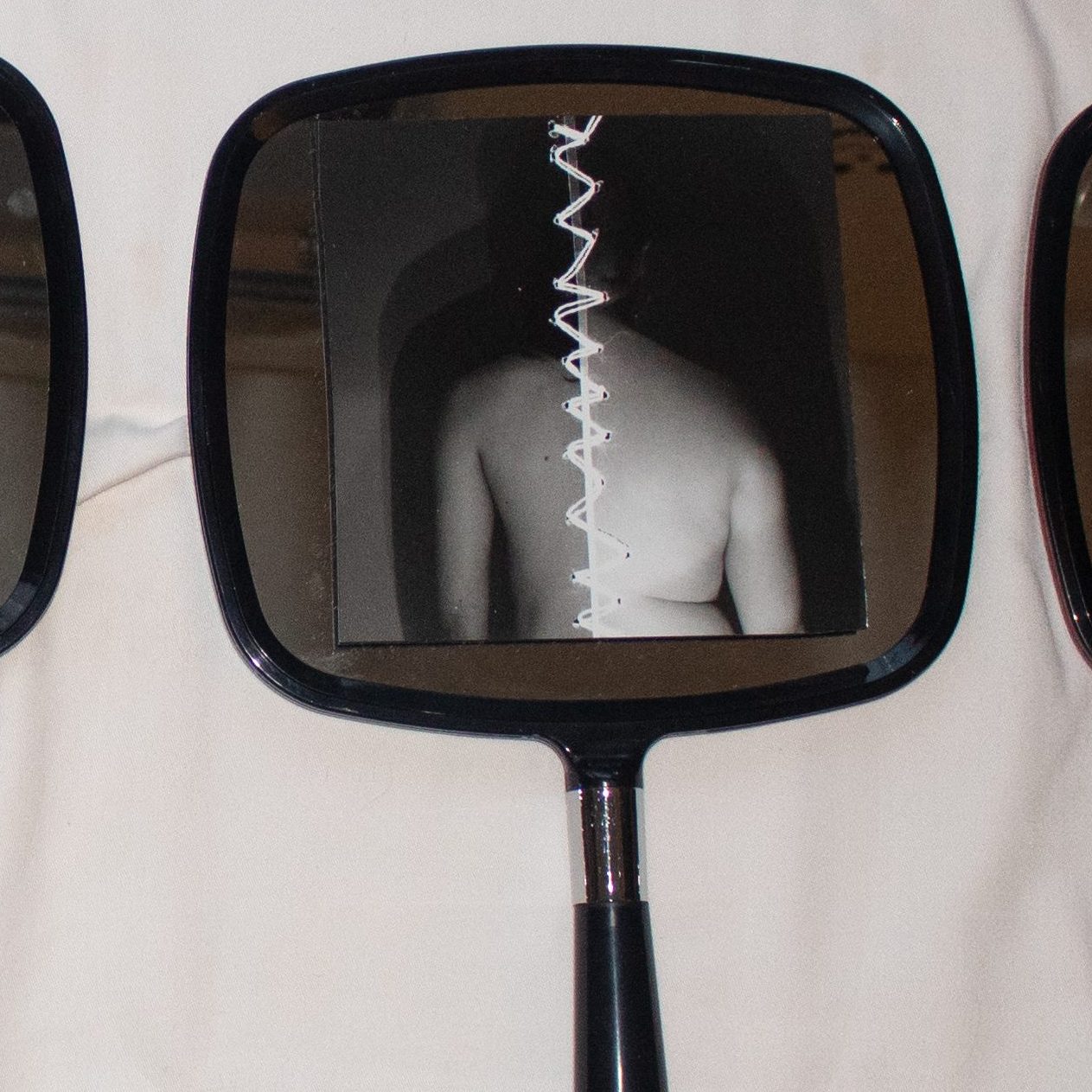 Five hand held mirrors lay on their back on a white sheet. On each mirror is a unique black and white image. The first image is a layered image of a person eating an apple, one layer is posed and proper, the other is smashed and messy. The proper layer is lighter in a ghostly manor. The second image in the series is of a naked curvy white female body from the side. Scratched out in the image are marks following along the woman's stretch marks. The third image is the body of a man without a shirt wearing dark pants. His hands are clasped together underneath a colostomy bag where a flower appears to be blooming. The fourth image is stitched together down the middle of a male and female body on either side of white stitches. The fifth and final image is of the back of a female body laying down as the curves integrate with hills and mountains making a sort of blended landscape within the image. 