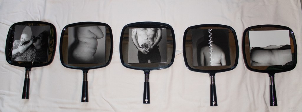 Five handheld mirrors lay on their back on a white sheet. On each mirror is a unique black and white image. The first image is a layered image of a person eating an apple, one layer is posed and proper, the other is smashed and messy. The proper layer is lighter in a ghostly manor. The second image in the series is of a naked curvy white female body from the side. Scratched out in the image are marks following along the woman's stretch marks. The third image is the body of a man without a shirt wearing dark pants. His hands are clasped together underneath a colostomy bag where a flower appears to be blooming. The fourth image is stitched together down the middle of a male and female body on either side of white stitches. The fifth and final image is of the back of a female body laying down as the curves integrate with hills and mountains making a sort of blended landscape within the image.  