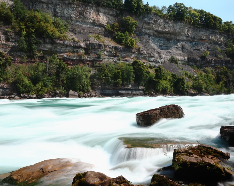 Flowing rapids rush around and above rocks, the water having an emerald green color. A cliff with trees is visible in the background.