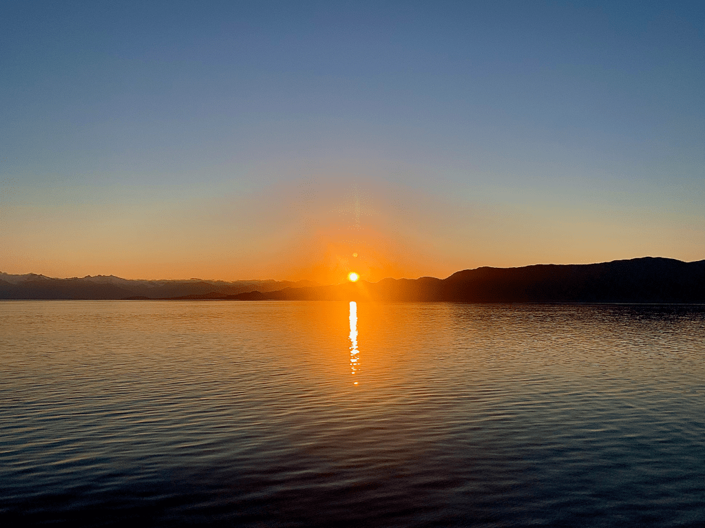 A horizontal photograph of the sunset, above a still body of water.  